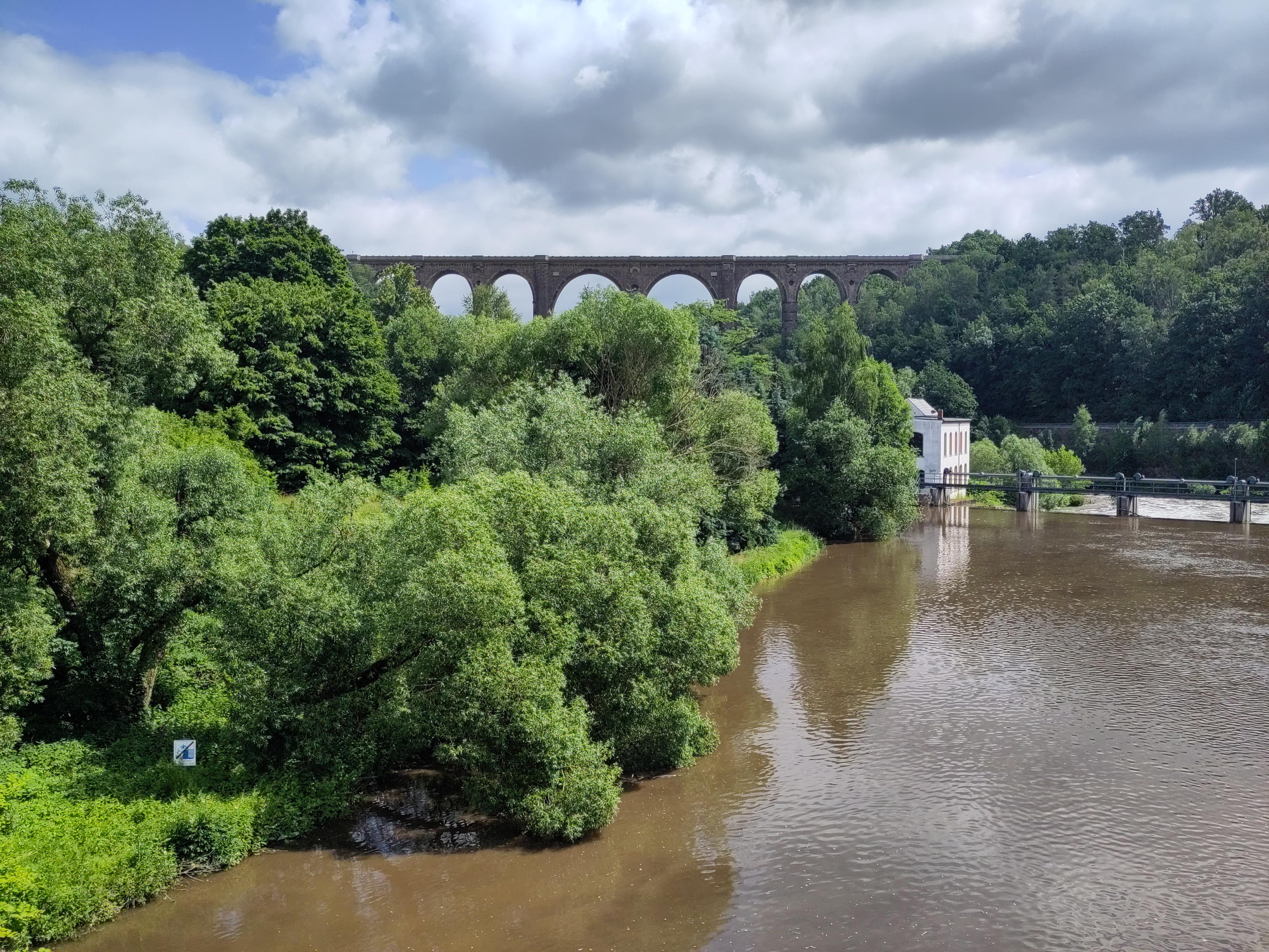 Ein brauner Fluß, ein Wehr mit einem Häuschen, im Hintergrund ist ein Viadukt zu sehen