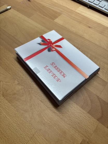 Ein kleiner flacher Karton mit der Aufschrift „Süßer Laptop“ und einer roten Geschenkschleife auf dessen Oberseite.