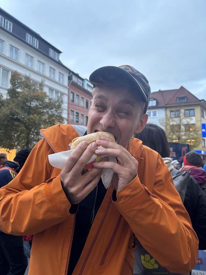 Ein Portrait von mir mit orangefarbener Regenjacke, grauer Cap während ich in ein Bratwurstbrötchen beiße. Im Hintergrund sind Protestierende (Omas gegen Rechts) vor der Würzburger Innenstadt.