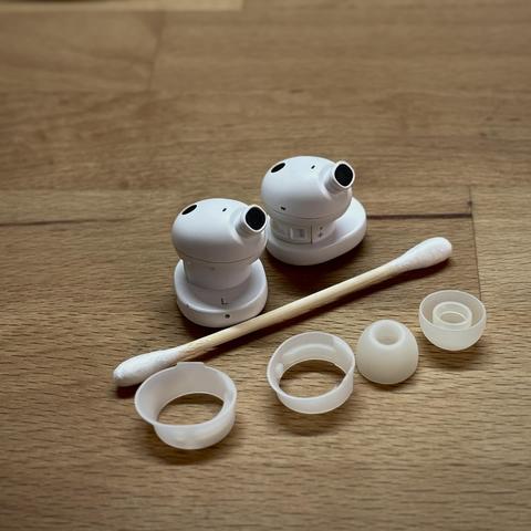 Weiße Fairbuds Kopfhörer diagonal mit ihren Silikonaufsätzen entlang eines Wattestäbchens aufgereiht. Sie liegen auf einer gewachsten Buchenholzplatte, die den Hintergrund bildet.