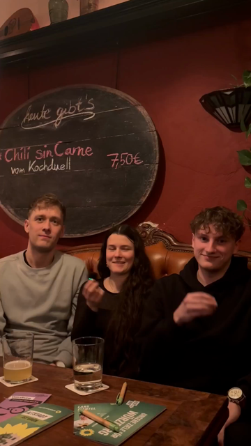 Zwei Männer und in der Mitte eine Frau sitzen zu dritt auf einer alten Ledercouch in der Bar Hanna Huckebein in Würzburg. Vor ihnen ist ein Holztisch mit Flyern und Getränken darauf. Das Video ist in Hochformat und sie sprechen nacheinander in die Kamera vor einer roten Wand mit einer großen ovalen Tafel, die Chili Sin Carne als Tagesgericht anpreist.