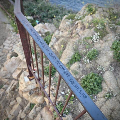 Ein rostiges Geländer an einem Felsen am Meer auf dessen Handlauf steht: „Ich bin so verliebt in Dich, wie Du in Autos!“