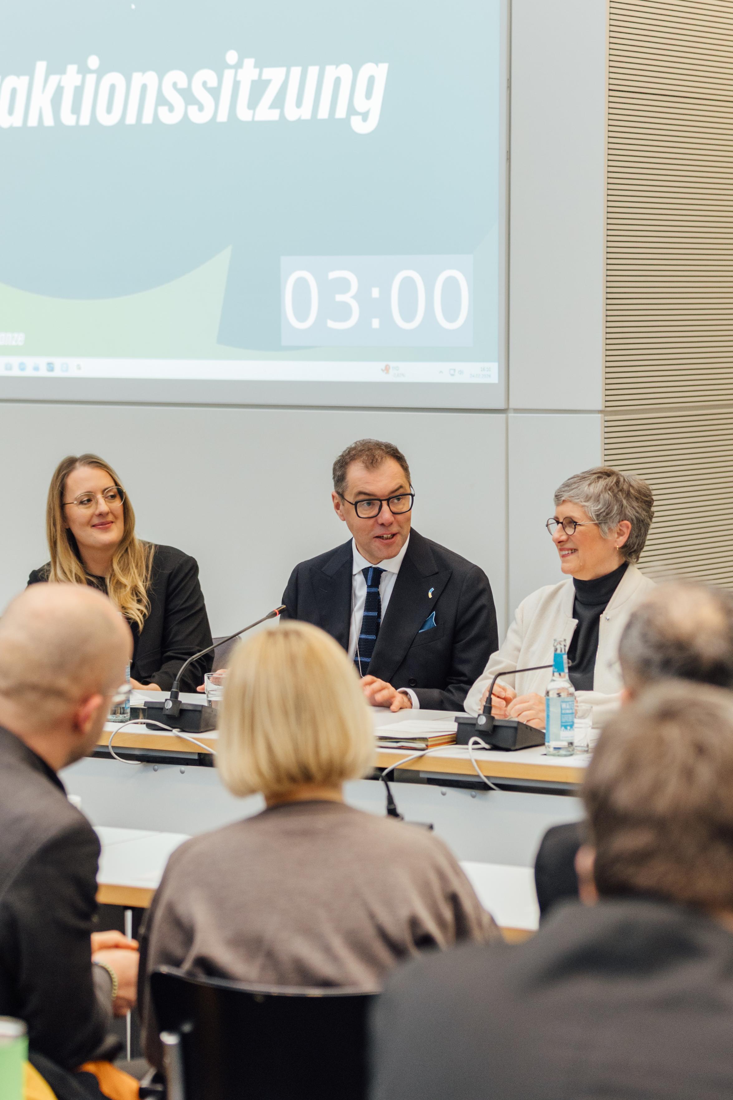 Der ukrainische Botschafter Oleksii Makeiev sitzt an einem Tisch in einem Saal. Neben ihm sitzen Britta Haßelmann und Katharina Dröge. Sie blicken sich an und lächeln.