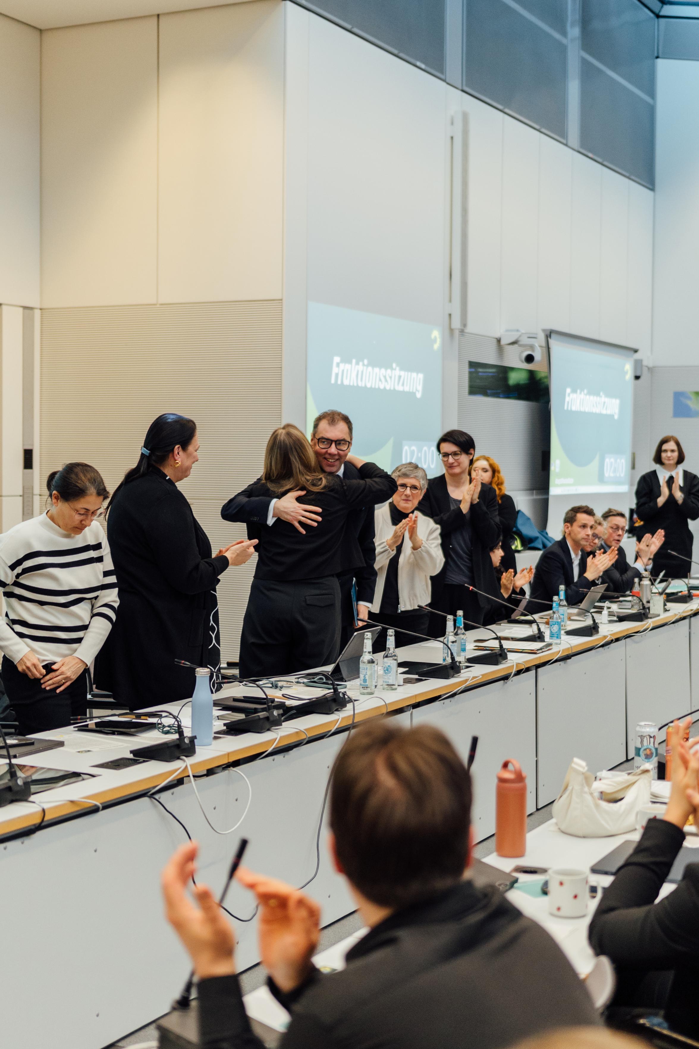 Abgeordnete der Grünen stehen in einem Raum an Tischen und klatschen in Richtung des Botschafters. Dieser umarmt Katharina Dröge, während andere Mitglieder des Vorstands der Grünen daneben stehen und klatschen.