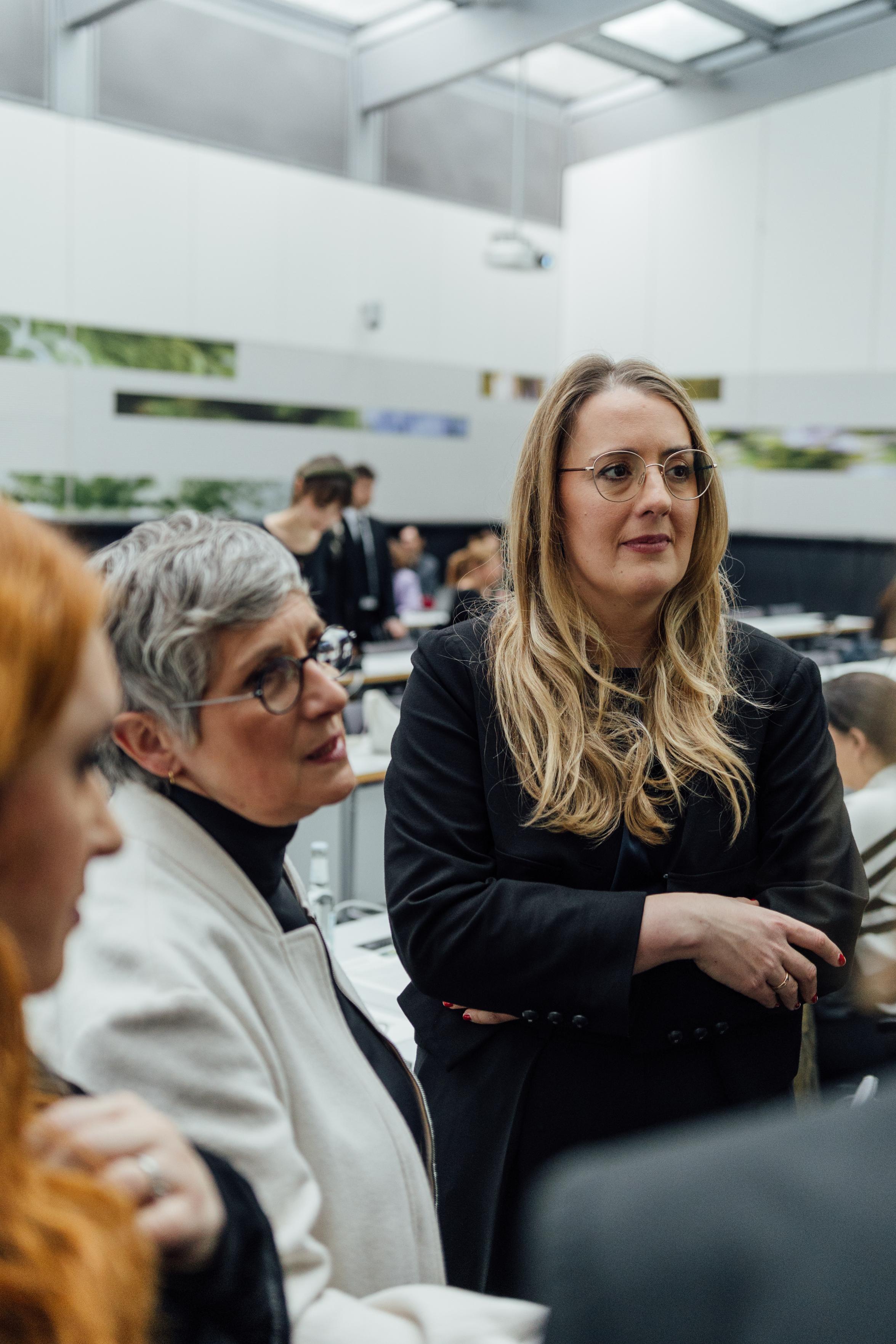 Katharina Dröge und Britta Haßelmann stehen in einer Gesprächsrunde und schauen auf den Botschafter, der im Anschnitt zu sehen ist. Sie reden angeregt.