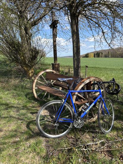 Ein blaues Rennrad lehnt an einer Bank aus alten, hölzernen Wagenrädern. Im Hintergrund sind Bäume, ein Flurkreuz und grüne Ackerflächen.